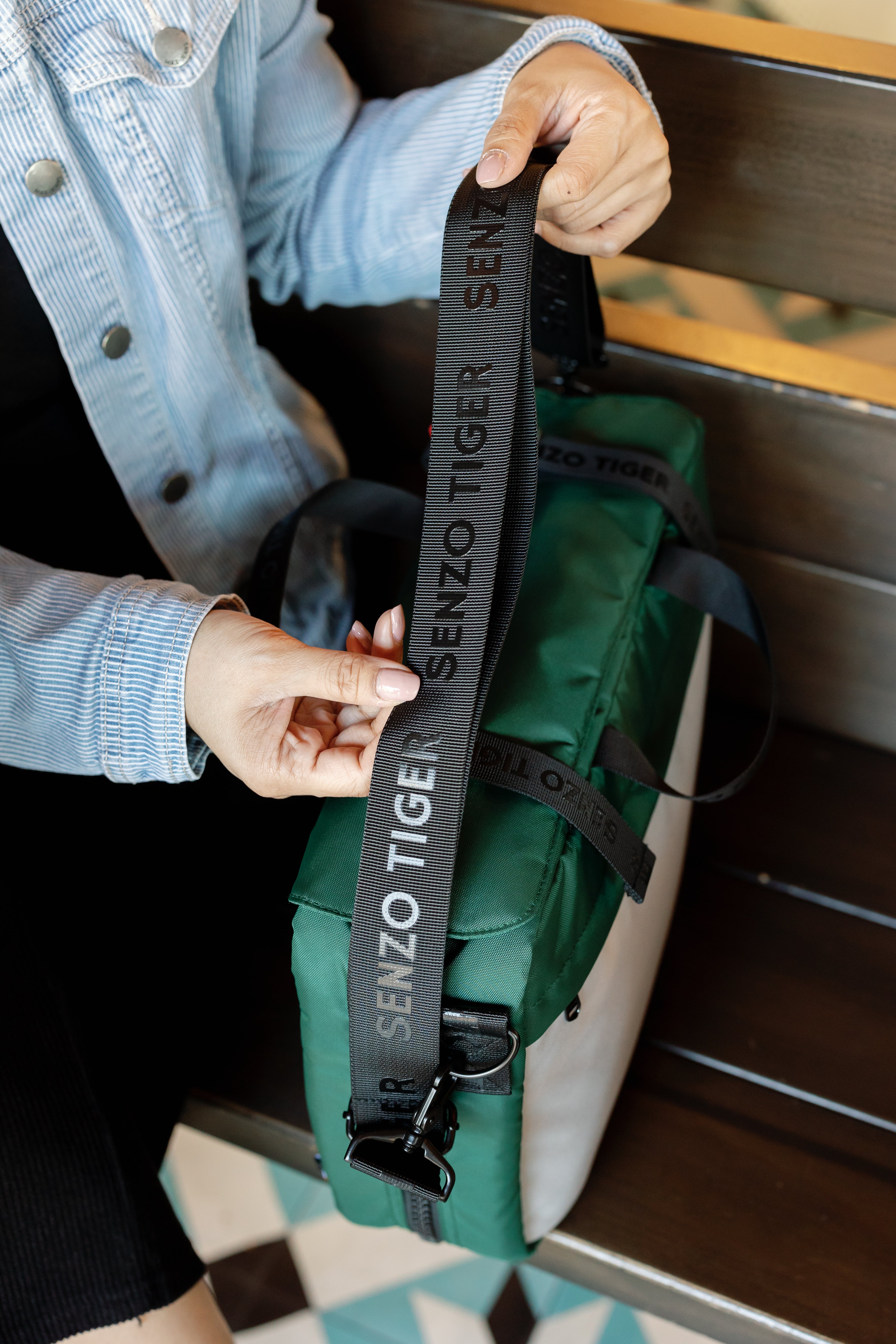 Woman holding up the shoulder strap of her green breifcase. The shoulder strap is branded with the words Senzo Tiger. 