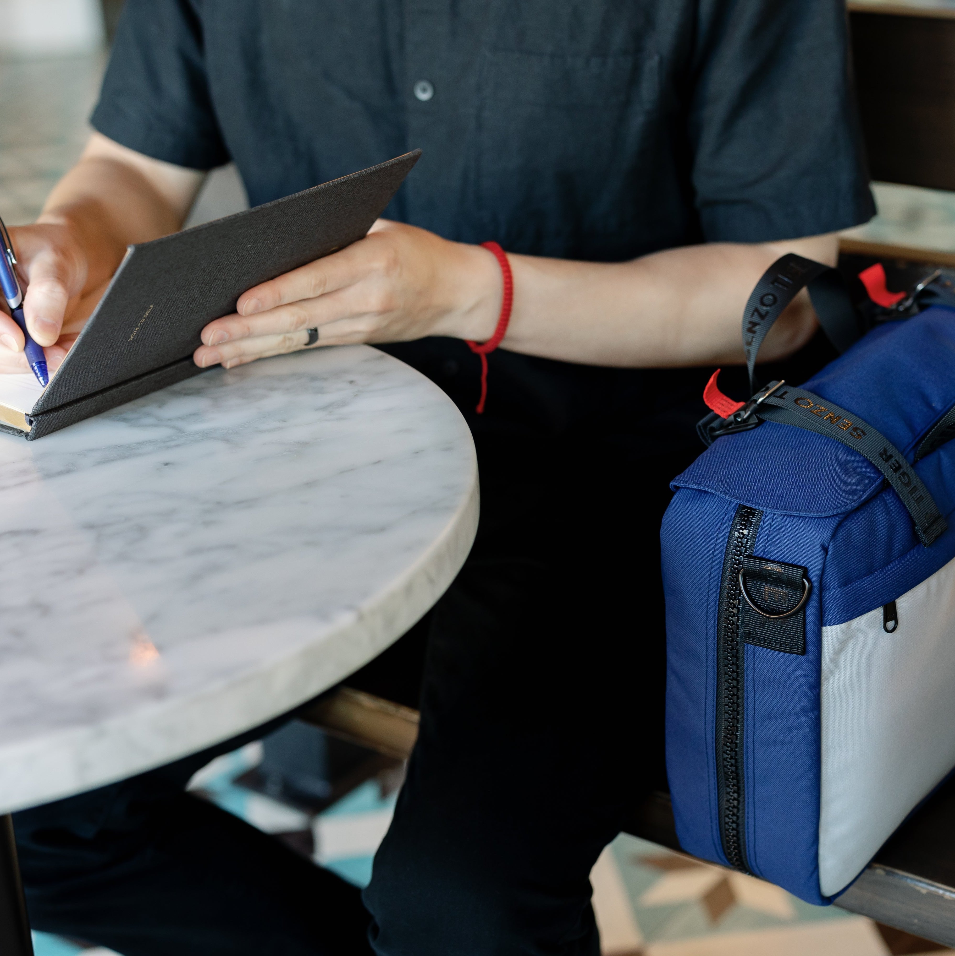 Man sitting at a coffee shop writing his notebook. His blue bag is beside him. 