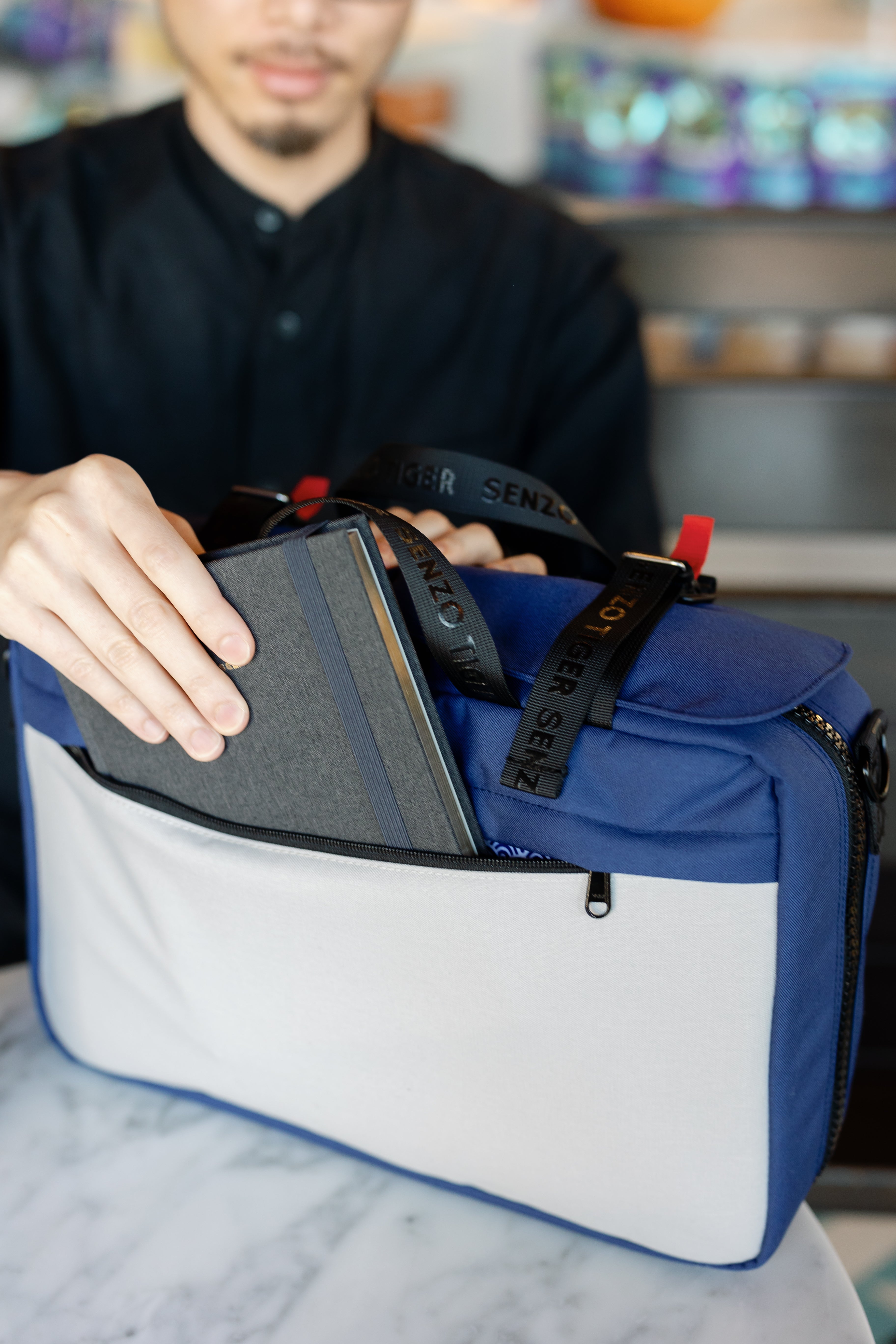 A man is pulling out a notebook out of the back pocket of his blue bag. You can see that there is plenty of space for a small notebook. 