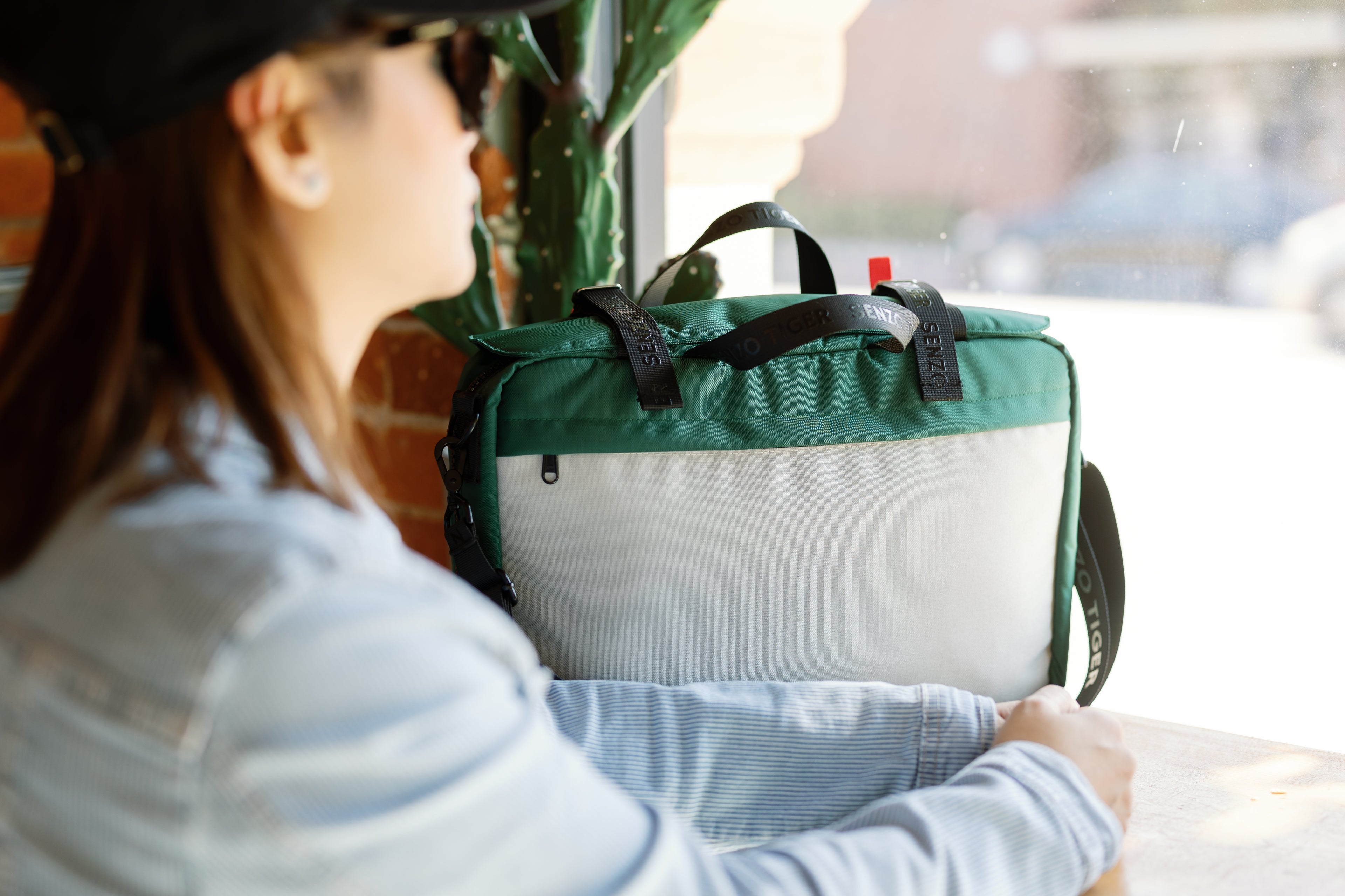 A woman in a demin jacke it looking out of a cafe window with her green bag beside her on a table counter. 