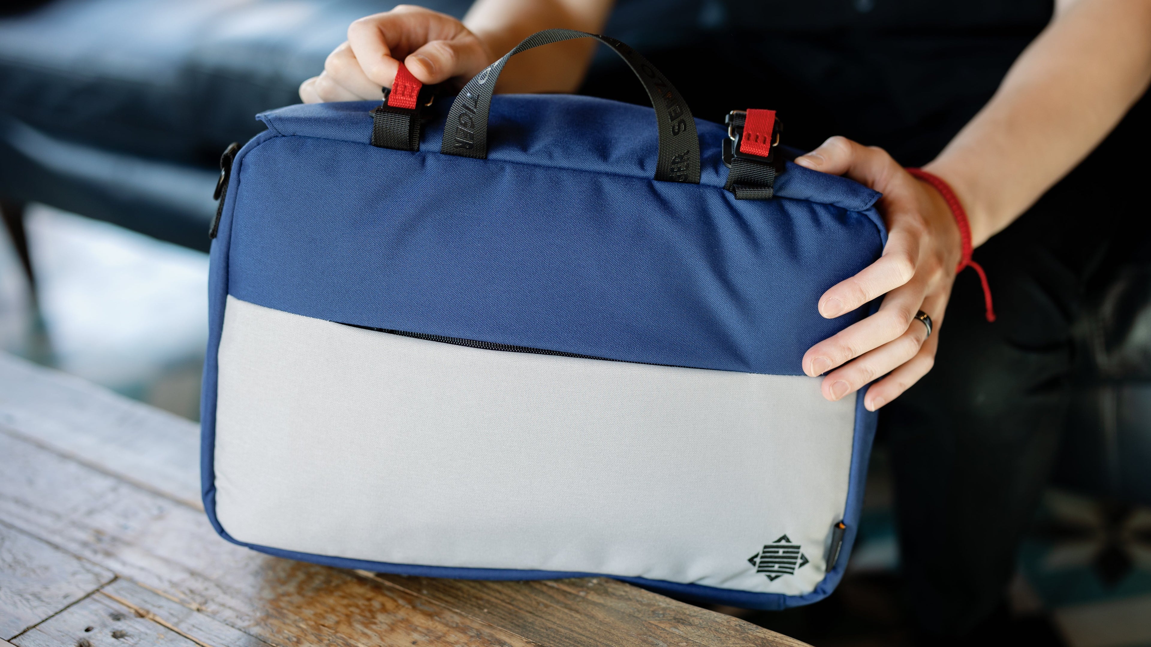 Man is placing the blue bag on a wooden table. His hand is puling on a magnetic Fidlock pull tab. 